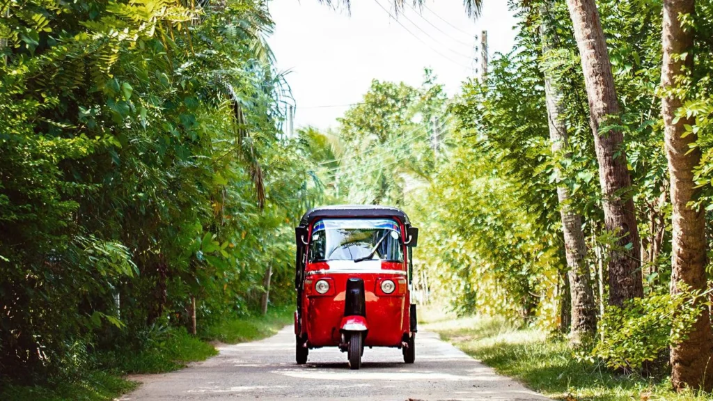 Red tuk-tuk travelling along a quiet tropical road surrounded by lush green trees in Sri Lanka.